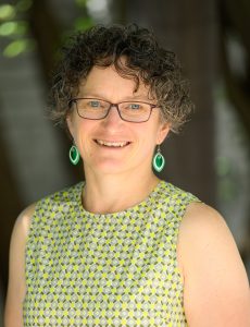 Photograph of Caroline Tiddy. Caroline is facing the camera and smiling. She is wearing a green, sleevless dress and green earrings. The background is blurred.